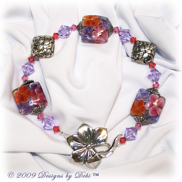 Designs by Debi Handmade Jewelry Aloha Collection Hawaiian Lei Bracelet featuring purple, pink and orange square aloha floral handmade lampwork beads, sterling lentil bead caps, bali antiqued silver embossed floral diamond-shaped pillows, swarovski crystal violet, rose and padparadscha bicones and a sterling silver large flower toggle clasp.